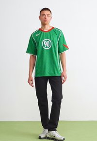 Groen sportshirt met rode accenten, witte bies en een cirkelvormig logo met "90" op de voorkant. Gecombineerd met zwarte broek en sportschoenen.