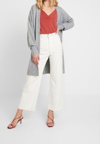 Grå lång kofta över en rostfärgad topp, kombinerad med vita vida byxor och röda sandaler med klackar och remmar. Släta texturer och rena linjer.