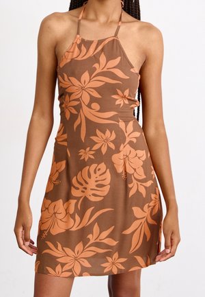 Femme portant une robe marron sans manches avec un imprimé floral orange et de fines bretelles dos nu, debout devant un fond uni.