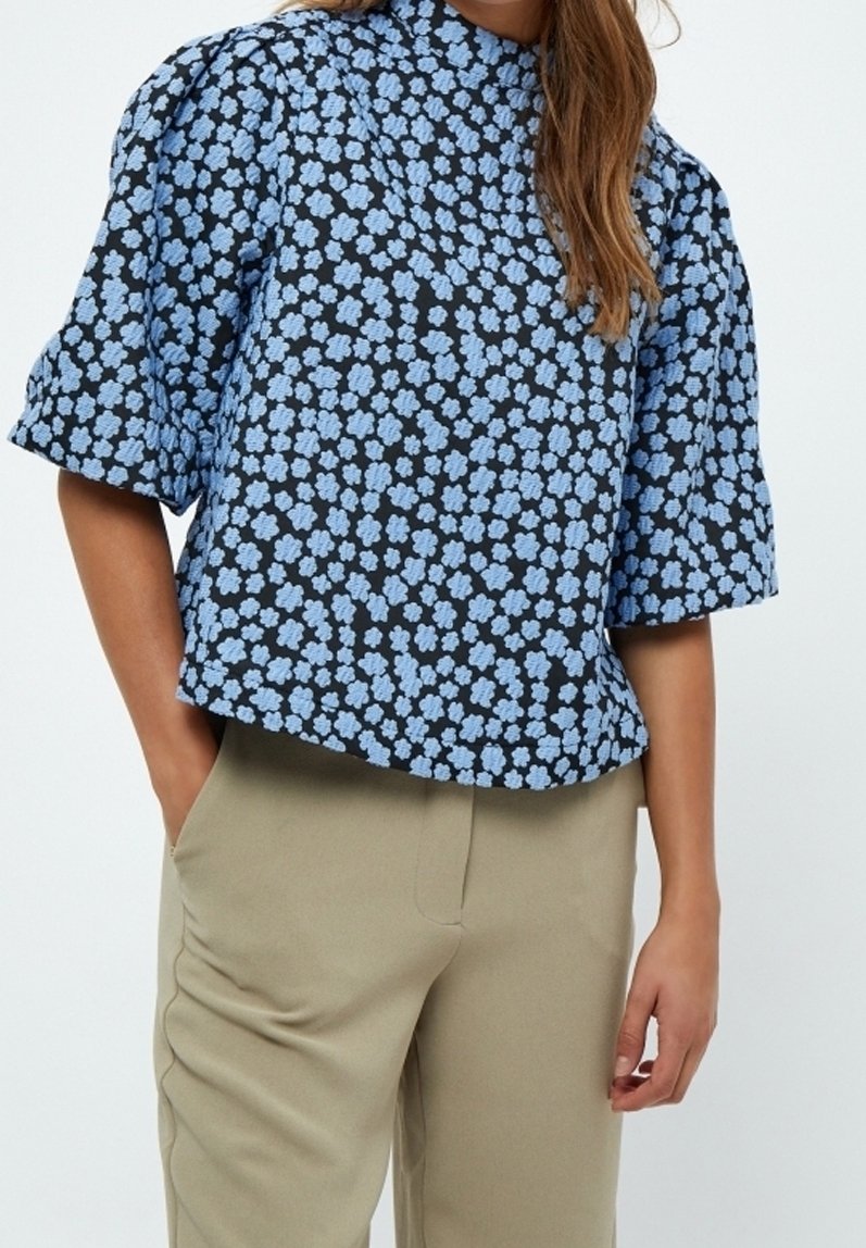 Femme portant un haut noir à manches courtes avec un motif floral bleu et un pantalon beige, une main dans la poche, sur un fond uni.