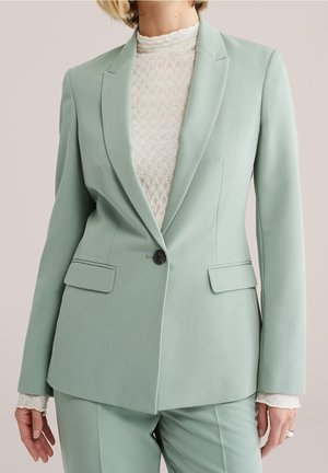 Femme portant un blazer vert clair cintré et un pantalon assorti sur un haut en dentelle blanche à col montant, visible du cou jusqu'à mi-cuisse.