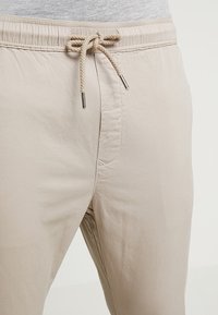 Beige lichtgewicht broek met een elastische tailleband, decoratieve trekkoorden en zijzakken; soepele stof met een subtiele glans.