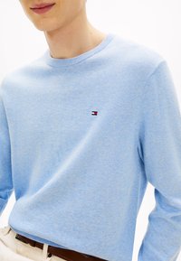 Svetlo modra pletena majica z okrolim izrezom, z majhnim logotipom v rdeči, beli in modri barvi na levi strani prsnega koša. Mehka tekstura in sproščen kroj.