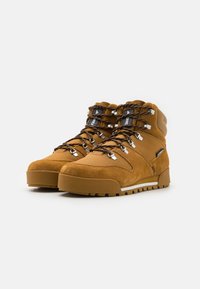 Brown suede hiking boots with black and yellow laces, metal eyelets, and a textured rubber sole. Features a cushioned ankle and logo accents.