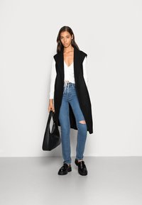 Casaco longo sem mangas preto sobre uma blusa branca, jeans skinny rasgados de azul claro e mocassins pretos. A modelo usa uma bolsa de ombro preta.