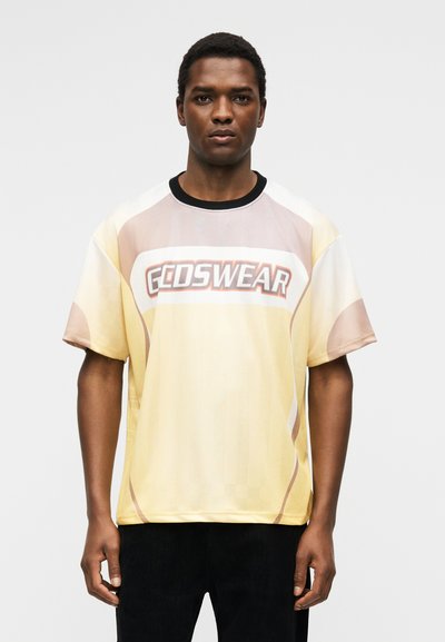 Άνδρας που φοράει κίτρινο και μπεζ κοντομάνικο πουκάμισο με το λογότυπο "GODSWEAR", στέκεται μπροστά σε απλό ανοιχτό γκρι φόντο.