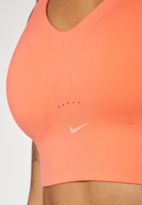 Nike sportski grudnjak u vibrantnom koraljnom tonu, napravljen od glatke tkanine. Ima zaobljeni dekolte, minimalno brendiranje i ventilacijske točkice iznad loga.