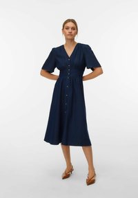 Kvinne iført en marineblå midikjole med korte ermer og knapper foran, kombinert med brune slingback-hæler med spiss tupp, stående med hendene på hoftene.