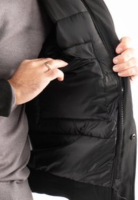 Veste noire à fermeture éclair sur le devant, présentant un intérieur lisse et matelassé ; poche proéminente visible. La texture du tissu semble douce et légère.