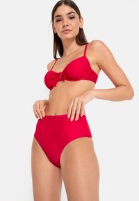Rdeč bikini komplet, ki vsebuje zavezani top in hlačke z višjim pasom. Gladko blago z nastavljivimi trakovi in polno pokritostjo.