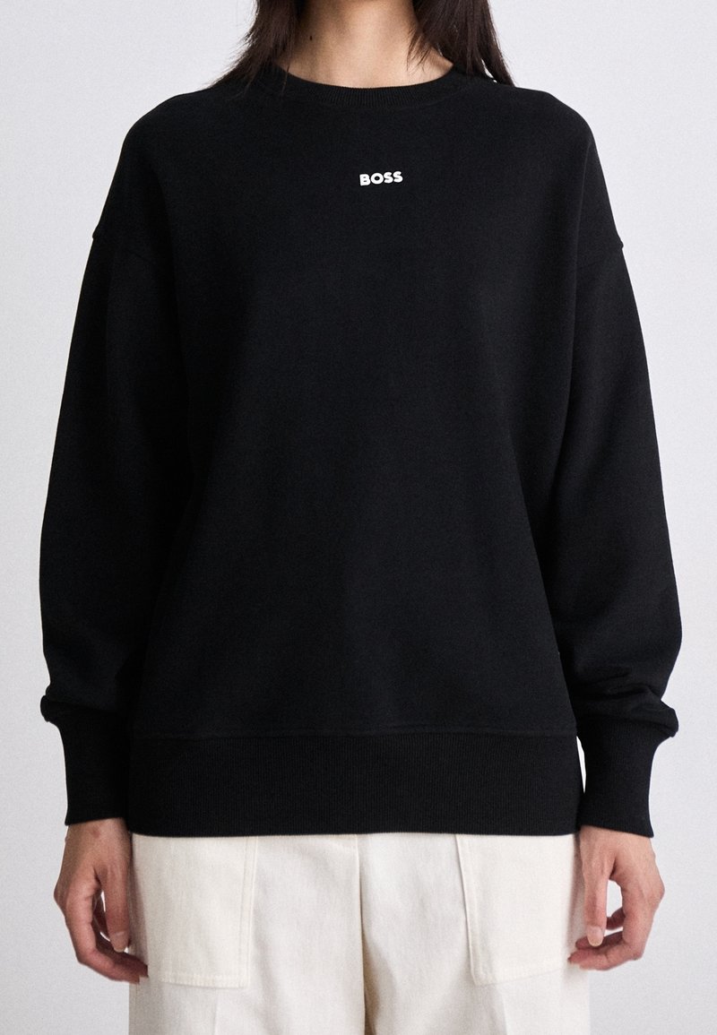 Personne portant un sweat-shirt noir à manches longues avec le mot « BOSS » en blanc sur la poitrine et un pantalon clair avec poches avant.