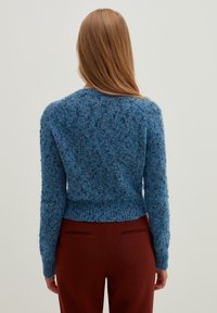 Kobieta z długimi jasnobrązowymi włosami, ubrana w niebieski sweter o teksturze i ciemnoczerwone spodnie, widziana od tyłu na jednolitym jasnym tle.