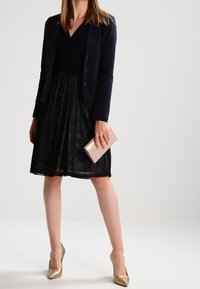 Musta mekko tekstuurikuvioilla, tummansininen bleiseri, kultaiset korot ja vaaleanpunainen clutch. Pehmeä kangas flare-hameen muotoilulla.