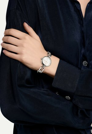 Montre argentée avec un cadran blanc rond et des index, dotée d'un bracelet texturé et d'un fermoir, portée sur une main contre un tissu sombre.