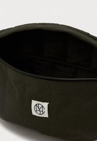 Μαύρος καπιτονέ σάκος με ανοιχτό φερμουάρ και μια μικρή λευκή τετράγωνη ετικέτα με μαύρο κυκλικό λογότυπο.