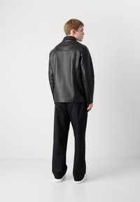 Chaqueta de cuero negro con una textura suave, corte entallado, costuras en los hombros y un collar puntiagudo. Llevada sobre pantalones oscuros y zapatos blancos.