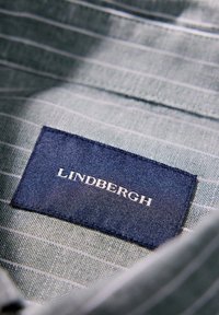 Tejido de rayas entrelazado en gris claro y blanco con una etiqueta azul marino que muestra "LINDBERGH" en texto blanco. Acabado texturizado y bordes cosidos.