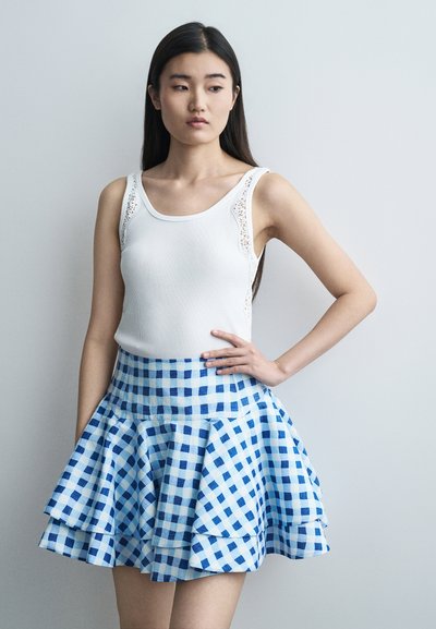 Jeune femme aux cheveux longs et foncés portant un haut blanc sans manches et une jupe à carreaux bleu et blanc, debout avec la main sur la hanche.