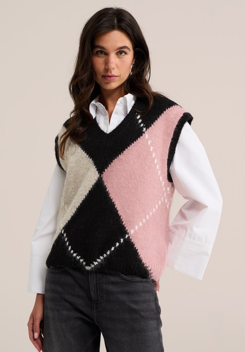 Femme aux cheveux foncés porte un gilet sans manches tricoté à motif losanges noir, rose et crème par-dessus une chemise blanche boutonnée et un jean foncé.