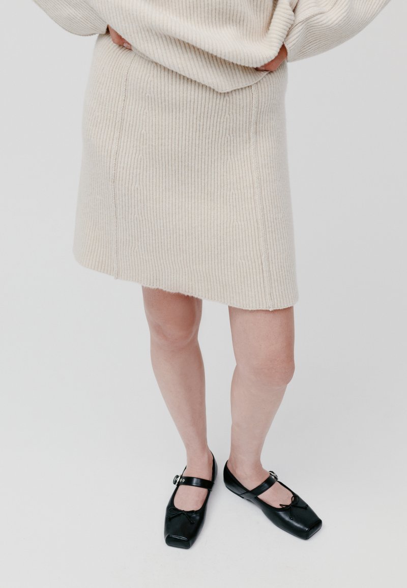 Hellbeige gerippter Strickrock mit geradem Schnitt, kombiniert mit schwarzen Lackleder-Schuhen mit quadratischer Zehenform, die über einen Riemen und eine dekorative Schleife verfügen.