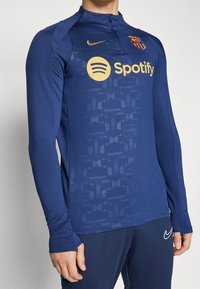 Lange mouwen, navy trainingsshirt van gladde stof. Beschikt over een ritskraag, Nike-logo en groot Spotify-logo in goud met textuurpatronen.