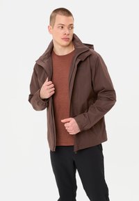 Bruine waterdichte jas met een capuchon, een gladde textuur, een rits aan de voorkant en verstelbare manchetten. Draagt over een maroon shirt met een ronde hals.