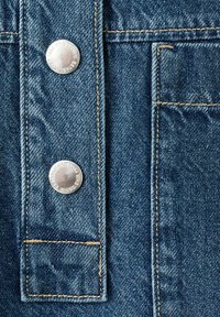 Tissu en denim en gros plan avec deux boutons métalliques argentés et des surpiqûres beiges visibles le long des bords des poches et de la bande de boutons.