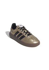 Zapatillas sintéticas doradas con tres franjas negras, cordones negros, parte superior texturizada y suela de goma. Marca en la lengüeta. Diseño de perfil bajo.