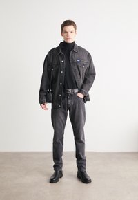 Black denim jacket over a black turtleneck, paired with black jeans and black boots. Model holds a small black bag, standing on a neutral floor.