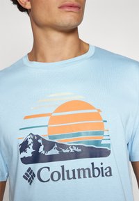 Lys blå t-skjorte med korte ermer som har et grafisk trykk med en solnedgang, fjell og "Columbia"-logoen foran. Myk bomullstoff, avslappet design.