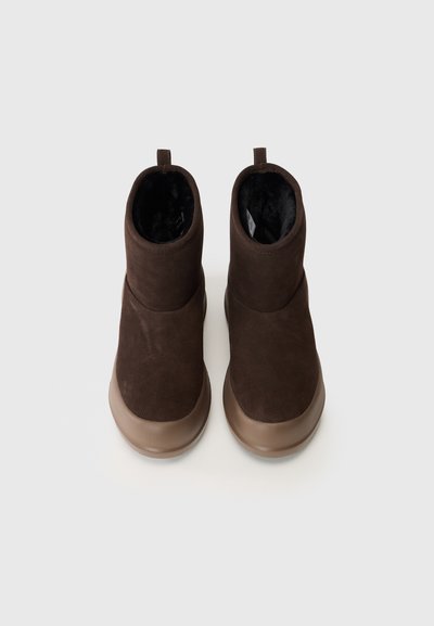 Ein Paar dunkelbraune Wildleder-Stiefeletten mit tanfarbenen Gummisohlen und schwarzem Plüschfutter, von oben auf einem hellen Hintergrund betrachtet.