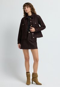 Giacca di jeans marrone con hardware dorato, indossata sopra un top scuro e abbinata a una gonna di jeans marrone coordinata. Stivali alla caviglia in suede beige.