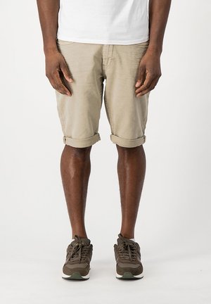 Homme portant un short beige retroussé, une chemise blanche et des baskets marron, debout devant un fond clair uni.