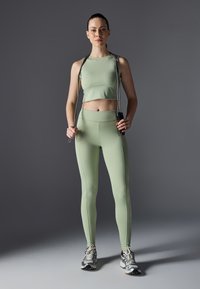 Conjunto deportivo de color verde claro que incluye una camiseta corta y leggings de talle alto, confeccionados en una tela suave. La modelo sostiene una cuerda para saltar frente a un fondo gris.