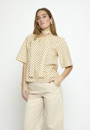 Mujer con blusa beige de manga corta con lunares y lazo en el cuello, y pantalones beige de pierna ancha, de pie con una mano en el bolsillo.