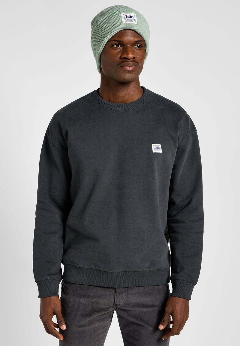 Ciemnoszara bluza z okrągłym dekoltem i ściągaczami, w połączeniu z jasnozielonym, prążkowanym czapką beanie z haftowaną naszywką logo.