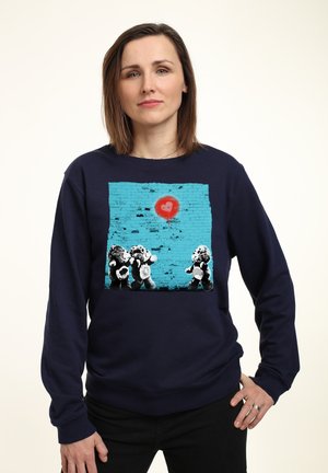 Mørkeblå sweatshirt med grafisk print af bamser på en teal murstensbaggrund med et rødt hjerte. 100% bomulds materiale, afslappet pasform, lange ærmer.