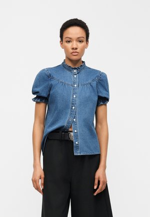Γυναίκα που φοράει μπλε κοντομάνικο τζιν πουκάμισο με γιακά με βολάν και μαύρο παντελόνι με φαρδιά ζώνη, πάνω σε απλό λευκό φόντο.