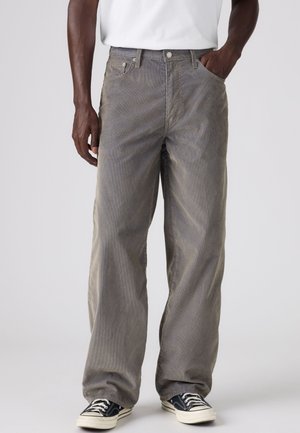 Homme portant un pantalon gris ample avec une chemise blanche rentrée, les mains dans les poches, et des baskets noires et blanches sur un fond uni.