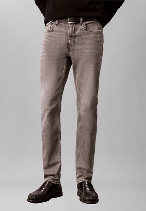 Mann trägt hellgraue Slim-Fit-Jeans, schwarzen Gürtel, dunkles Oberteil und schwarze Lederschuhe, steht vor einem schlichten Hintergrund.