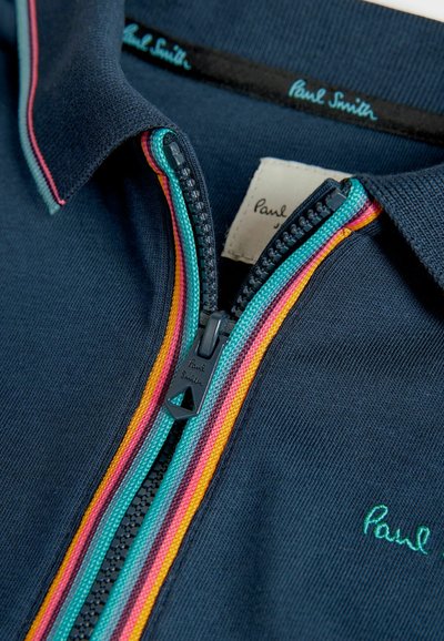 Mornarsko polo majico z ovratnikom na zadrgo. Ima večbarvne zadrge v roza, oranžni in turkizni barvi. Vključuje logotip "Paul Smith" v zeleni barvi.