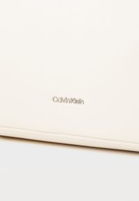 Fehér bőr táska sima textúrával, az elején ezüstszínű "Calvin Klein" logóval. Minimalista dizájn és diszkrét díszítések.