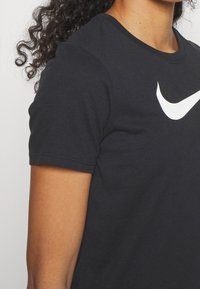 Camiseta negra de algodón con cuello redondo y mangas cortas. Presenta un prominente logo blanco de Nike en forma de swoosh en el frente.