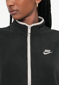 Črna flis jakna na zadrgo z visokim ovratnikom in sivo obrobo. Ima bel Nike logotip na prednjem delu. Gladka tekstura.