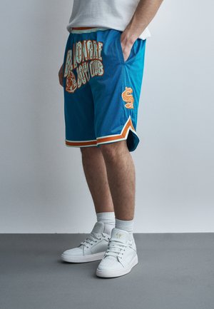 Persona che indossa pantaloncini da basket blu con bordi arancioni e bianchi, con la scritta "Billionaire Boys Club", scarpe da ginnastica alte bianche, in piedi su un pavimento grigio.