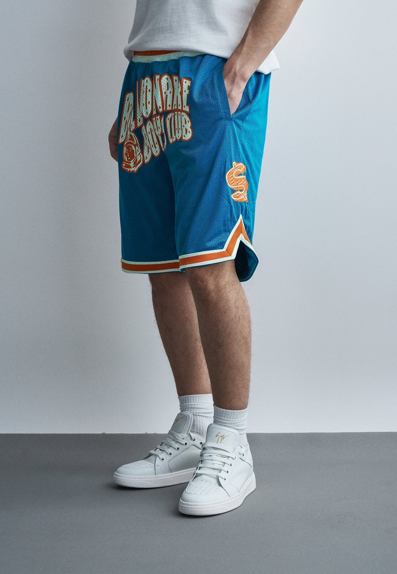 Személy kék kosárlabdás rövidnadrágot visel narancs és fehér szegéllyel, amelyen "Billionaire Boys Club" felirat található, fehér magas szárú edzőcipőben, szürke padlón állva.