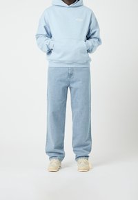 Person wearing light blue hoodie, light blue jeans, and beige sneakers, standing with hands in hoodie pocket on white background.