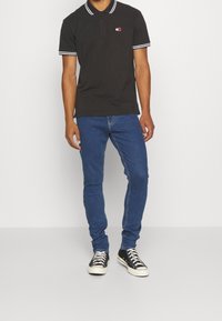 Svart pikétröja med vit krage och ärmskarvar, broderad logotyp i röd och vit, i kombination med blå slim-fit jeans och svarta sneakers.