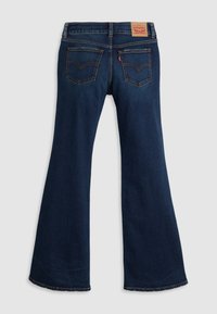 Jean en denim bleu foncé avec une jambe évasée, deux poches arrière avec coutures et un patch de marque en cuir sur la ceinture.