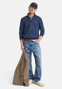 Marineblauer Reißverschluss-Pullover, hellgraues Hemd darunter, blaue Jeans, hält eine braun karierten Jacke, trägt eine marineblaue Mütze und weiße Sneakers.